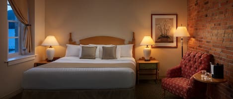 A hotel room with a large bed, two bedside tables with lamps, a red armchair, and a brick wall.