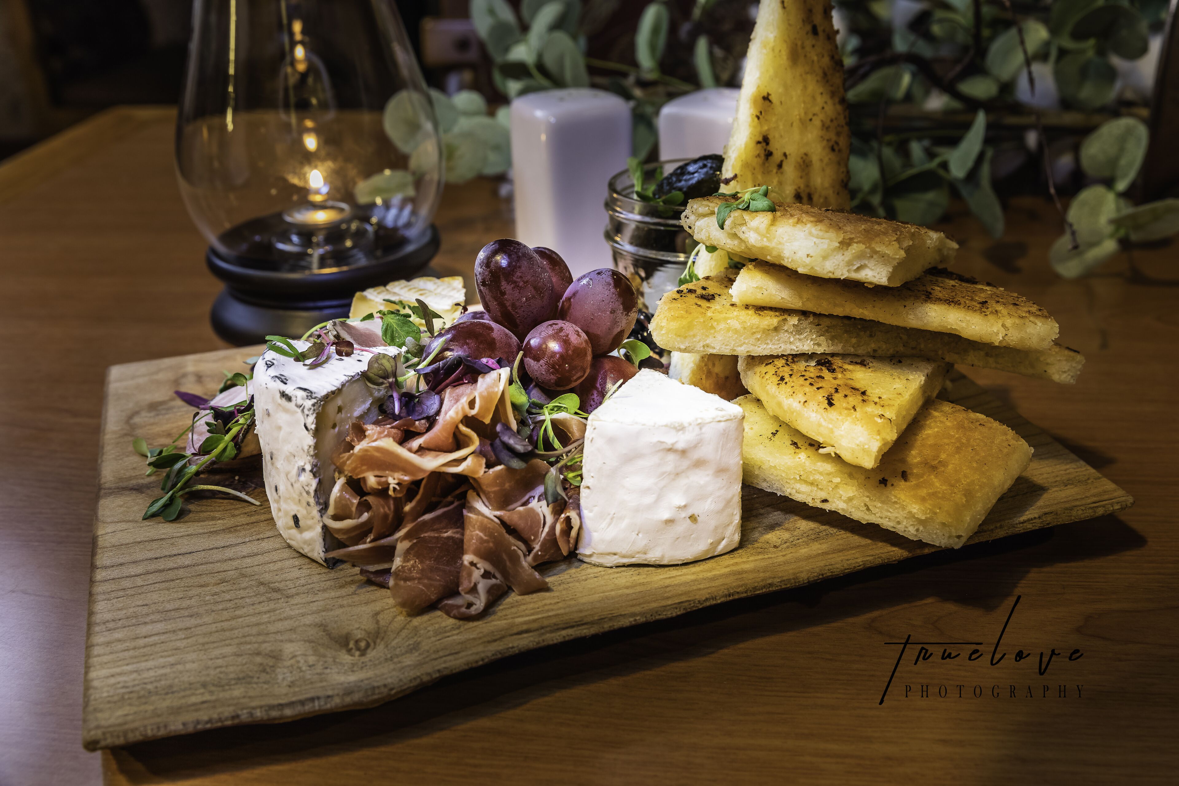 Une planche en bois sur laquelle sont disposés divers aliments, notamment du fromage, de la charcuterie et du pain, accompagnée d'un porte-bougie en verre et de quelques éléments de verdure en arrière-plan.