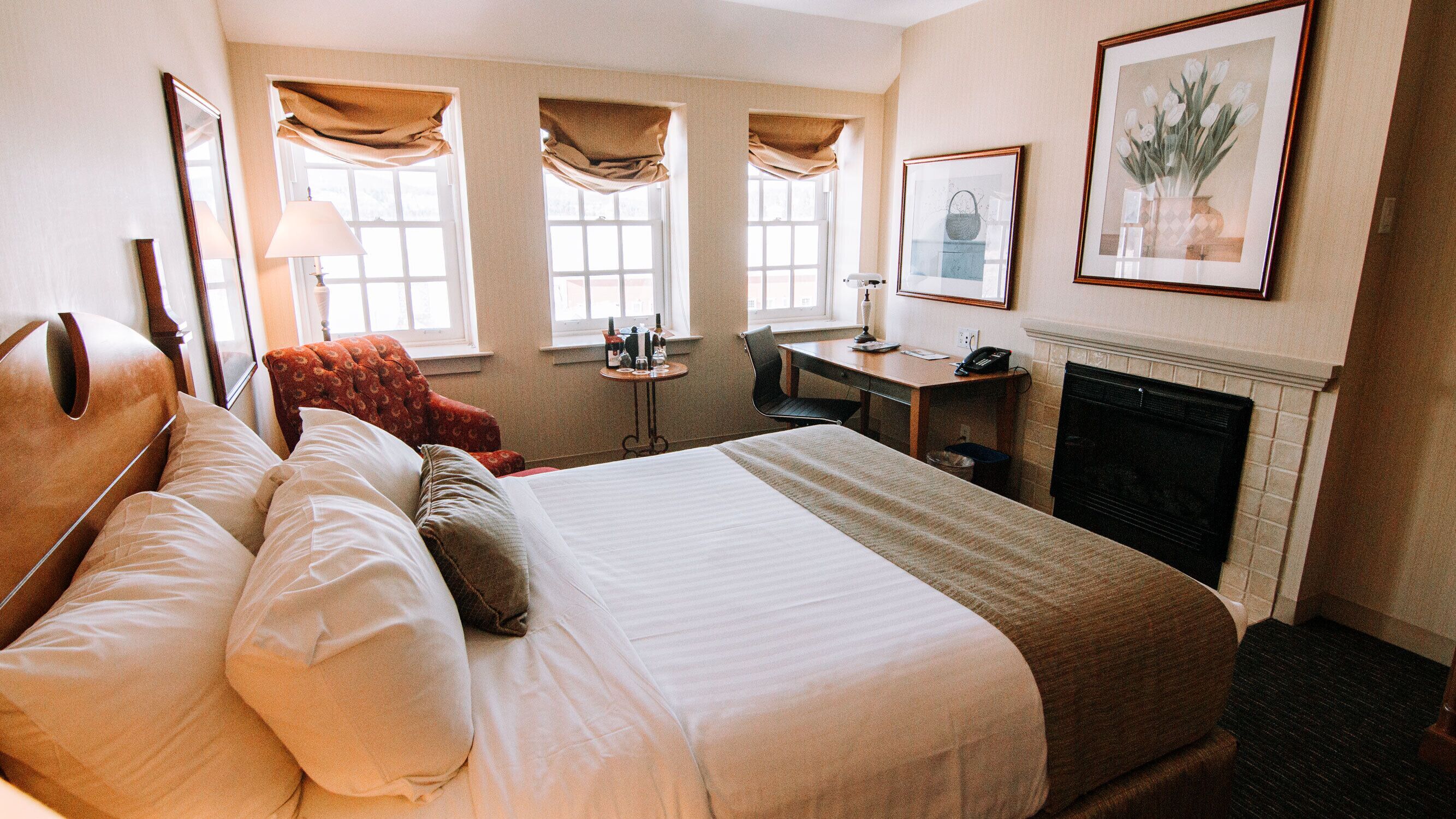 A hotel room with a large bed, a desk, a chair, a fireplace, and two windows with curtains.