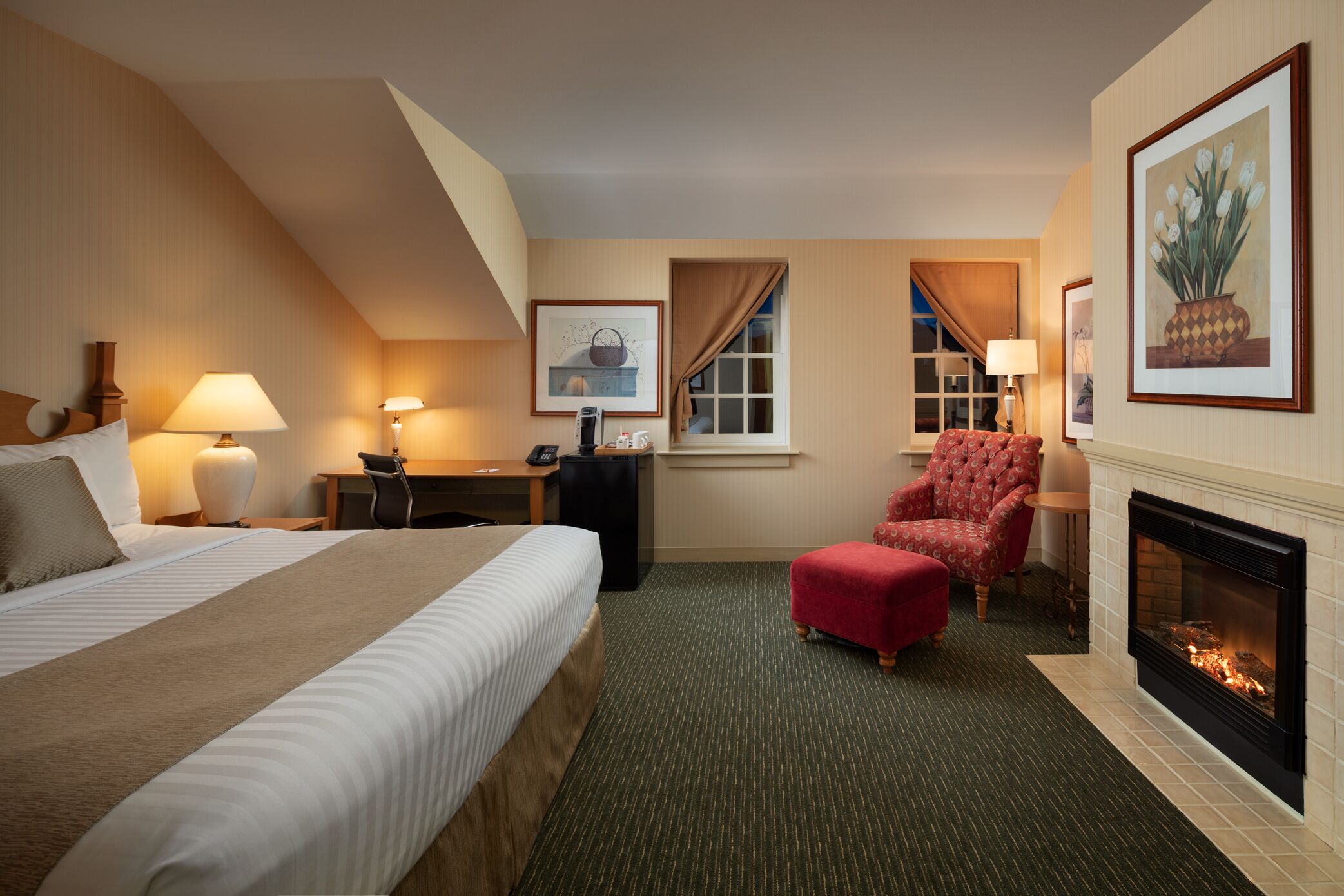 A hotel room with a large bed, a desk, a red armchair, a fireplace, and a window with curtains.