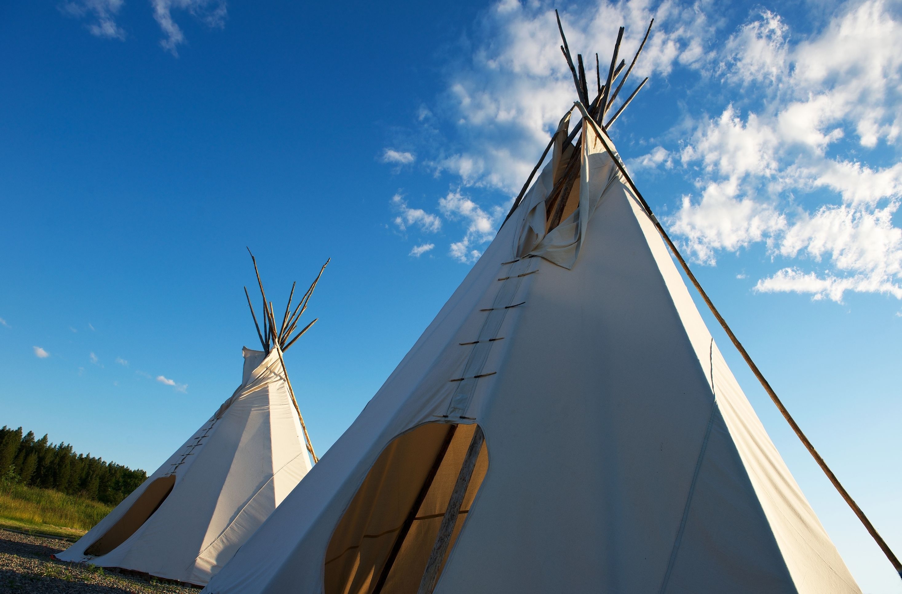 Two white teepees with wooden poles, set against a clear blue sky with scattered clouds.
