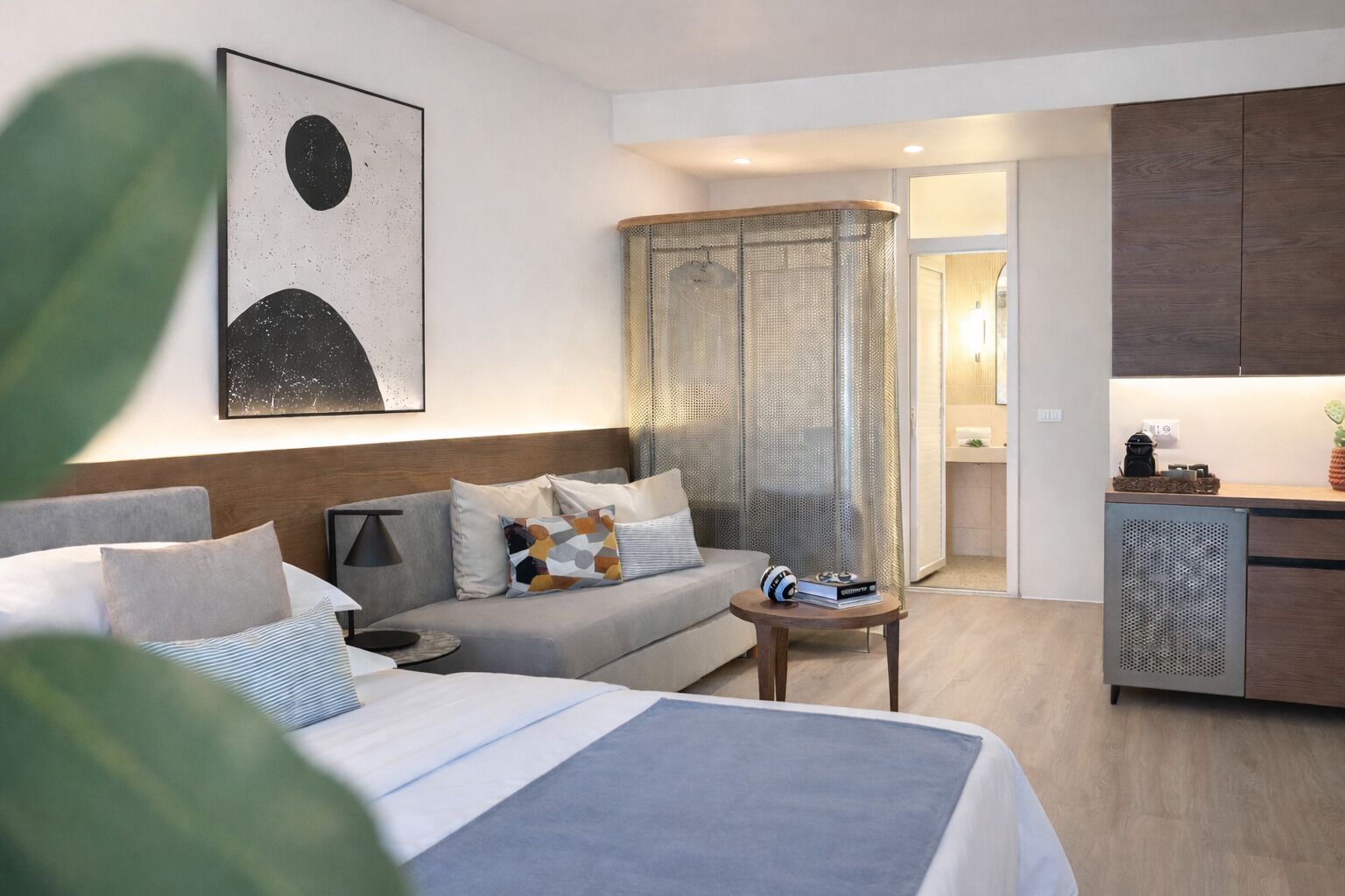 A modern hotel room with a bed, a sofa, a small table, and a kitchenette.