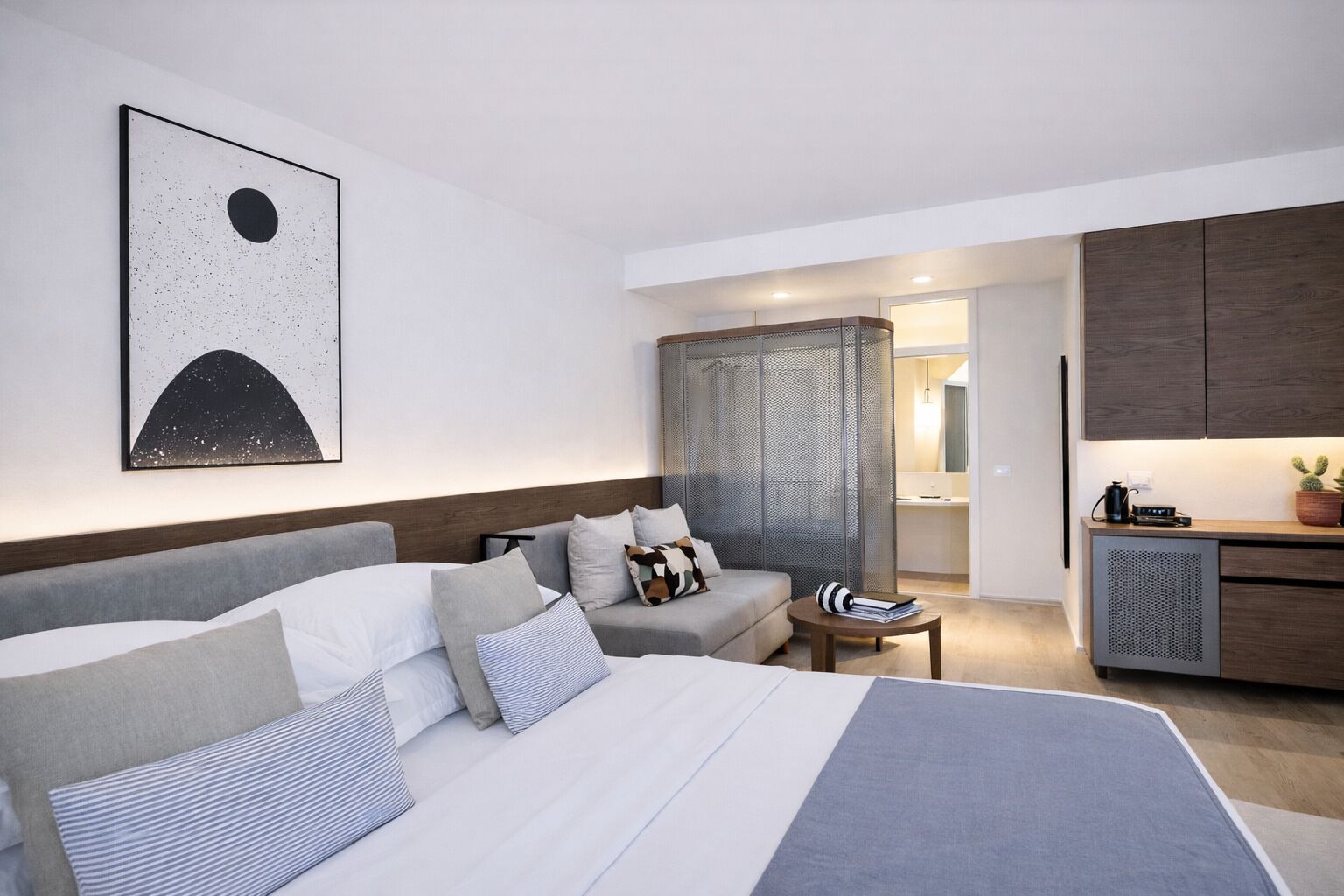 A modern hotel room with a large bed, a sofa, a small table, a kitchenette, and a decorative wall piece.