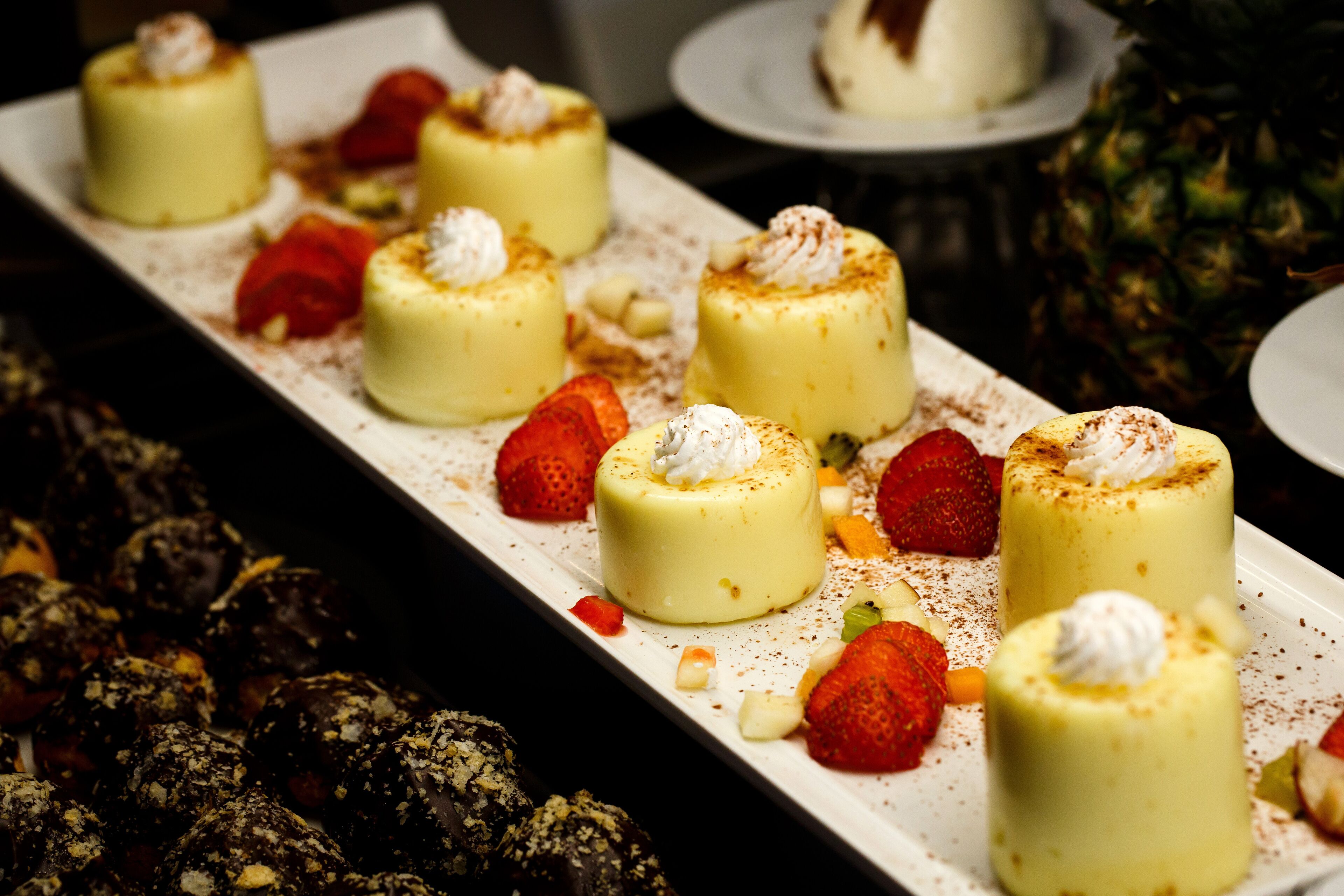 Een dessertpresentatie met kleine, ronde, gele desserts, afgetopperd met slagroom en bestrooid met cacaopoeder, vergezeld van aardbeien en ander fruit.