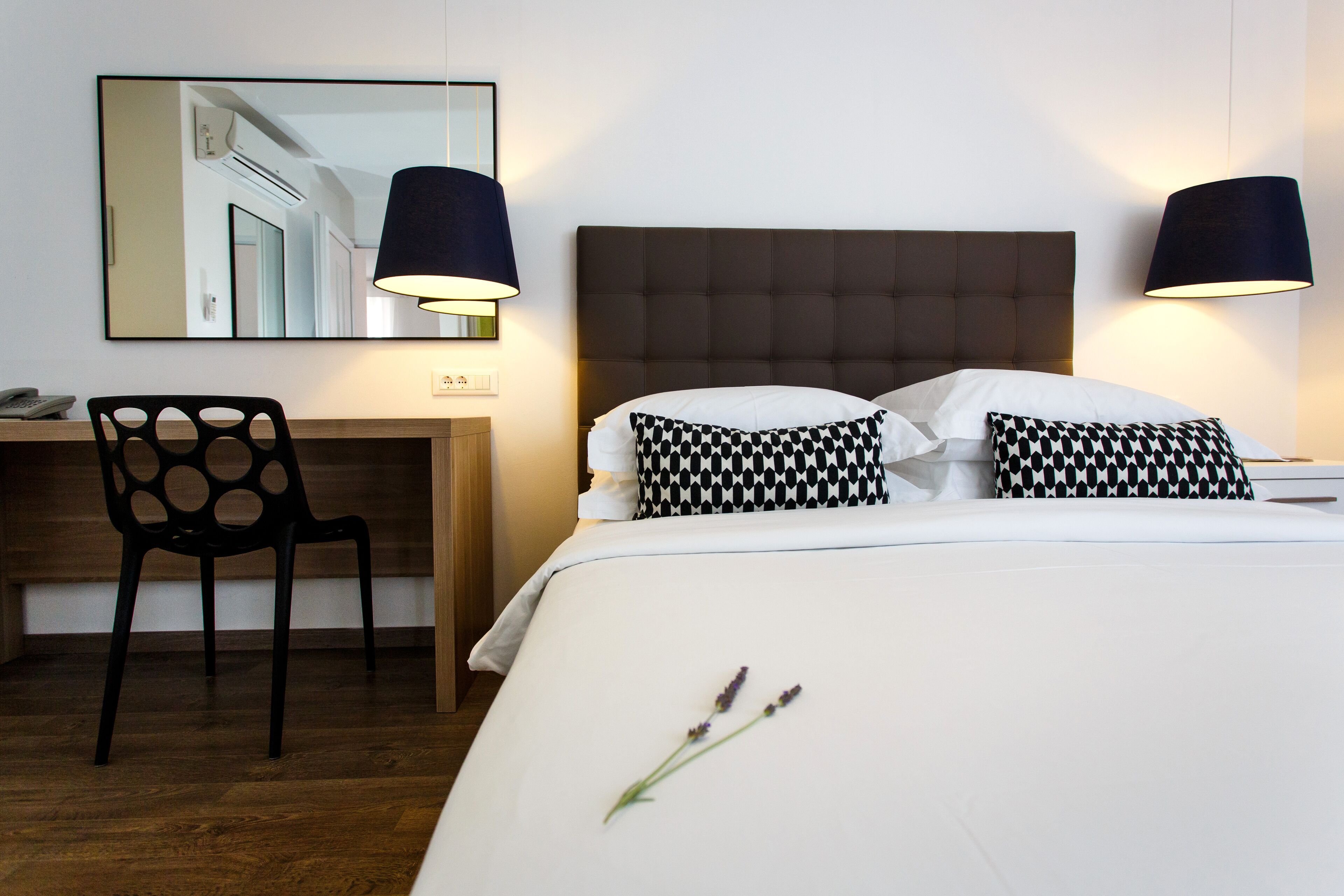 Una camera d'albergo con un letto, due lampade nere, una scrivania con una sedia e uno specchio a parete.
