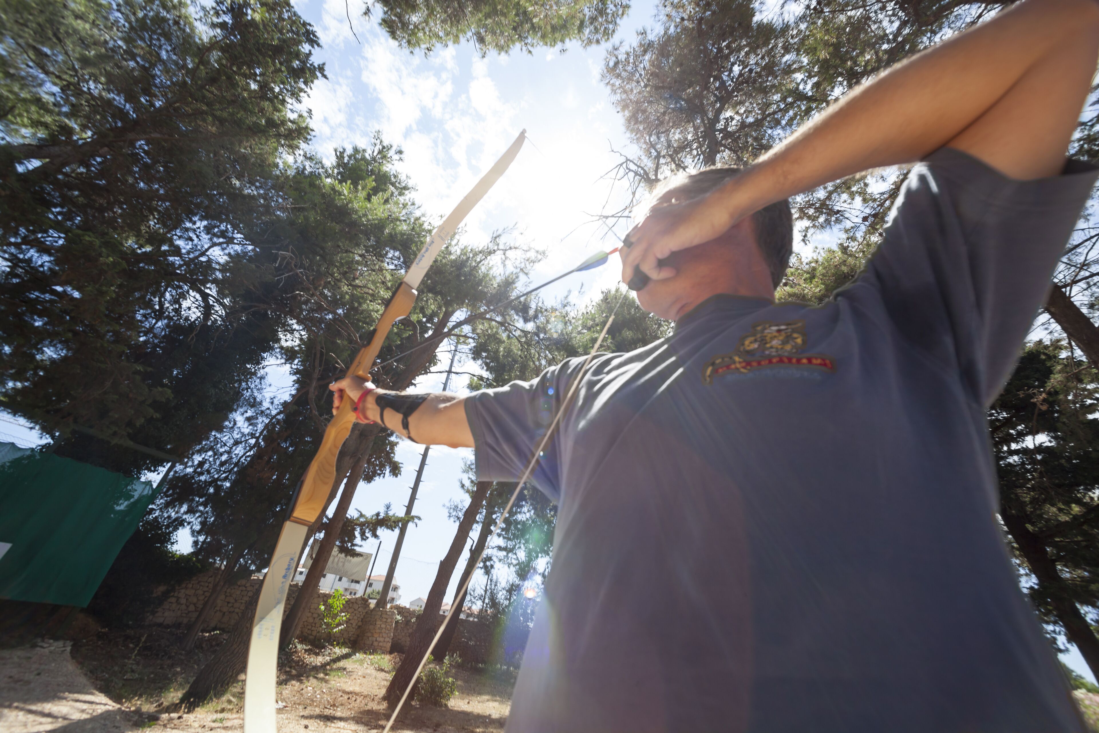 A person holding a bow and arrow outdoors with trees in the background.