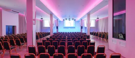 A conference room with rows of chairs, a stage with a large screen, and large windows.