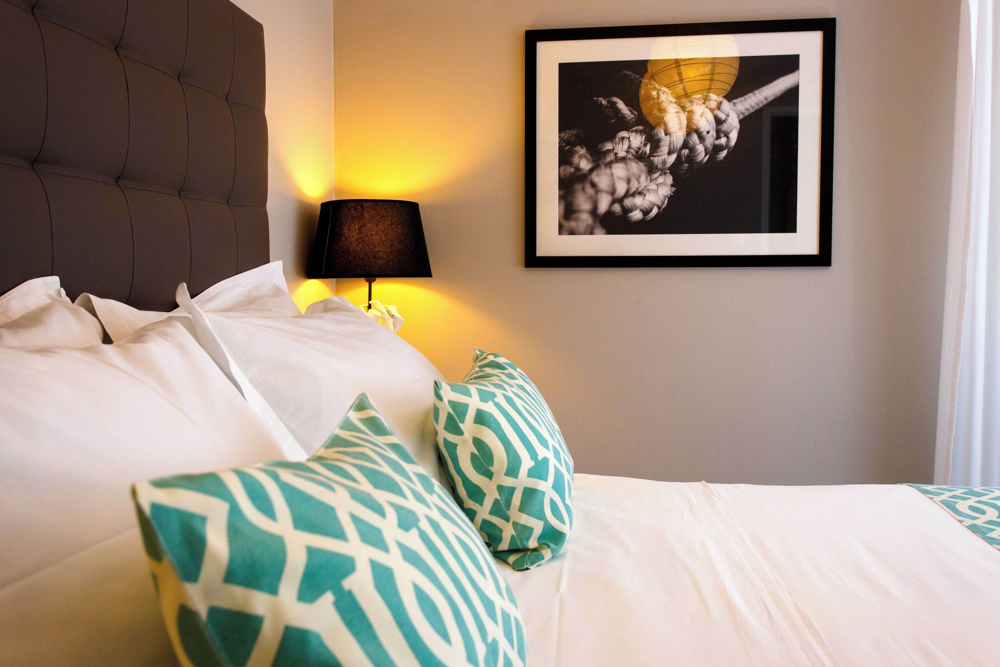 A neatly made bed with white bedding and green patterned pillows, a wall-mounted lamp, and a framed picture on the wall.