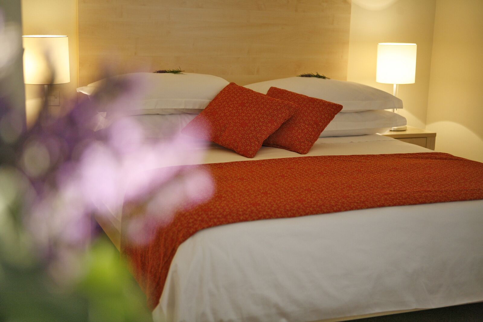 Una cama bien hecha con ropa de cama blanca y dos almohadas rojas, una manta roja y dos lámparas de noche en una mesita.