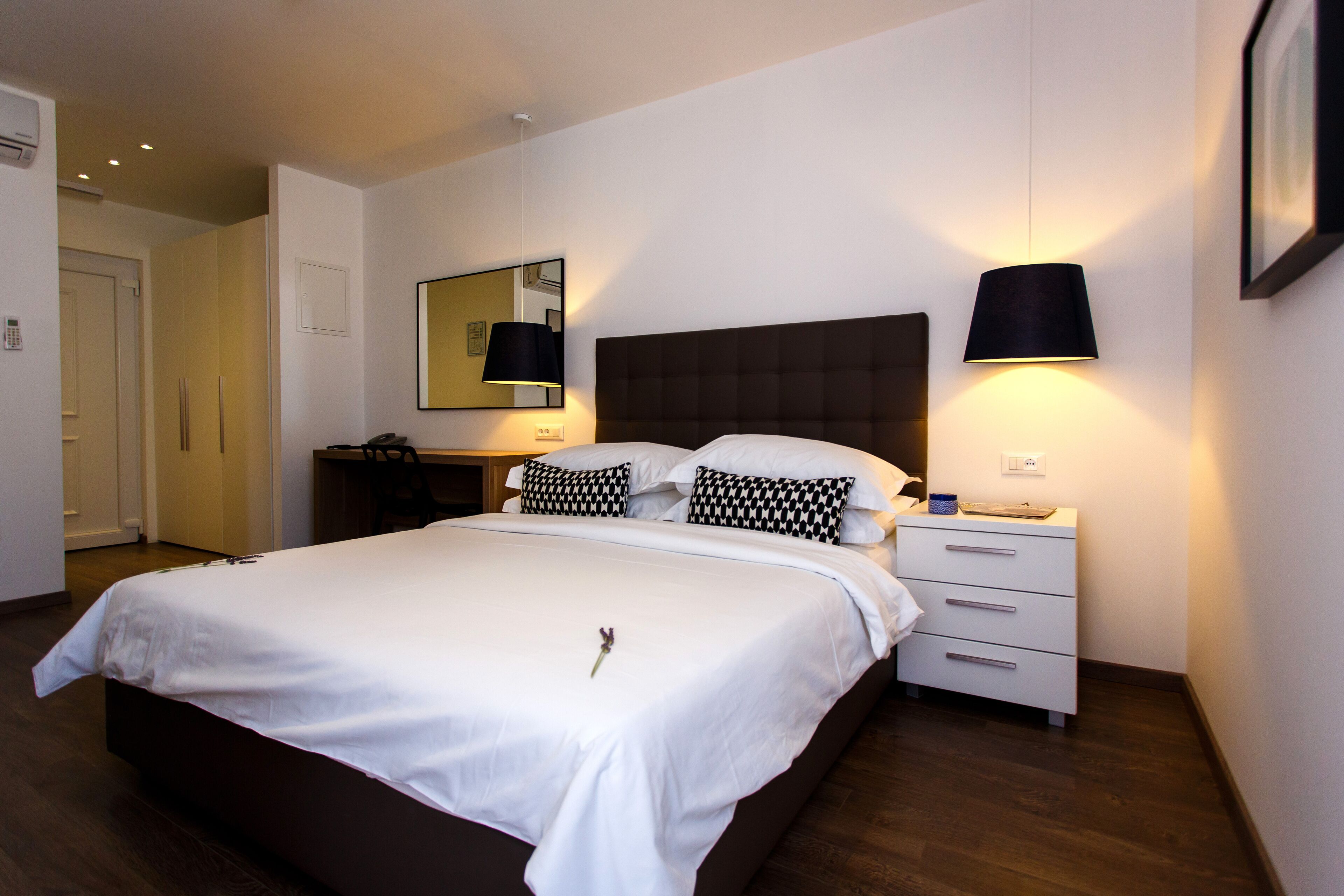 Een net opgemaakt bed met witte lakens en kussens in zwart-wit patroon, een nachtkastje met een lamp, en een klein dressoir.