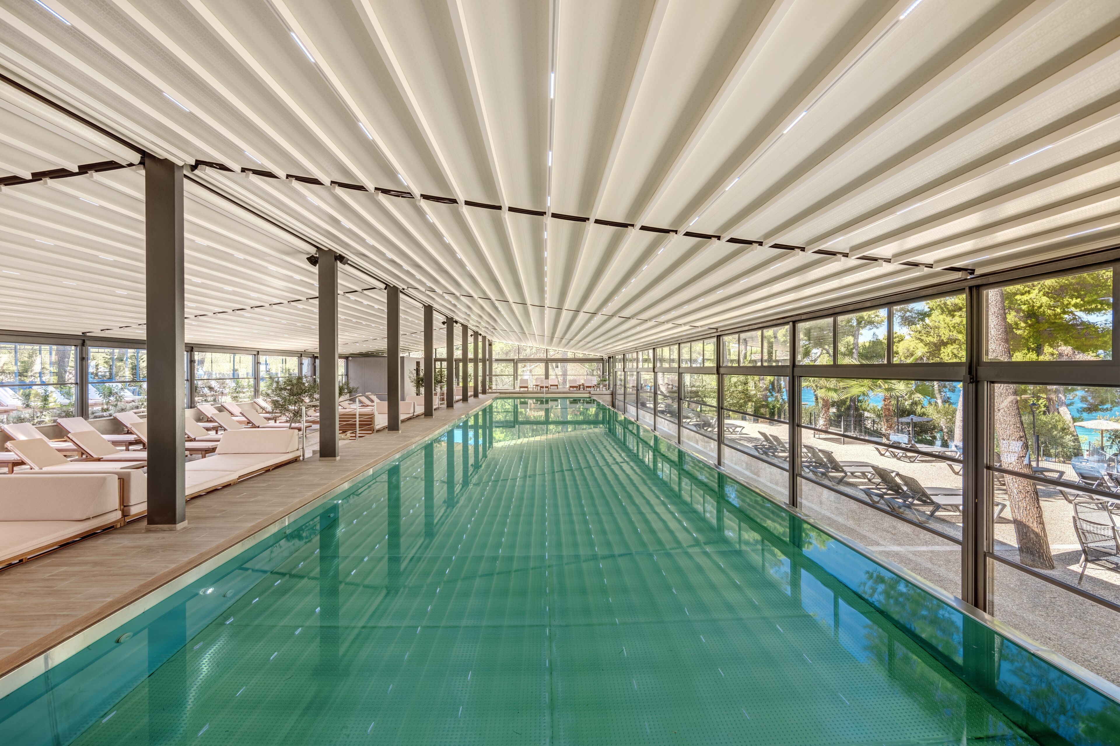 A large indoor swimming pool with a glass barrier, lounge chairs, and a view of the outside.