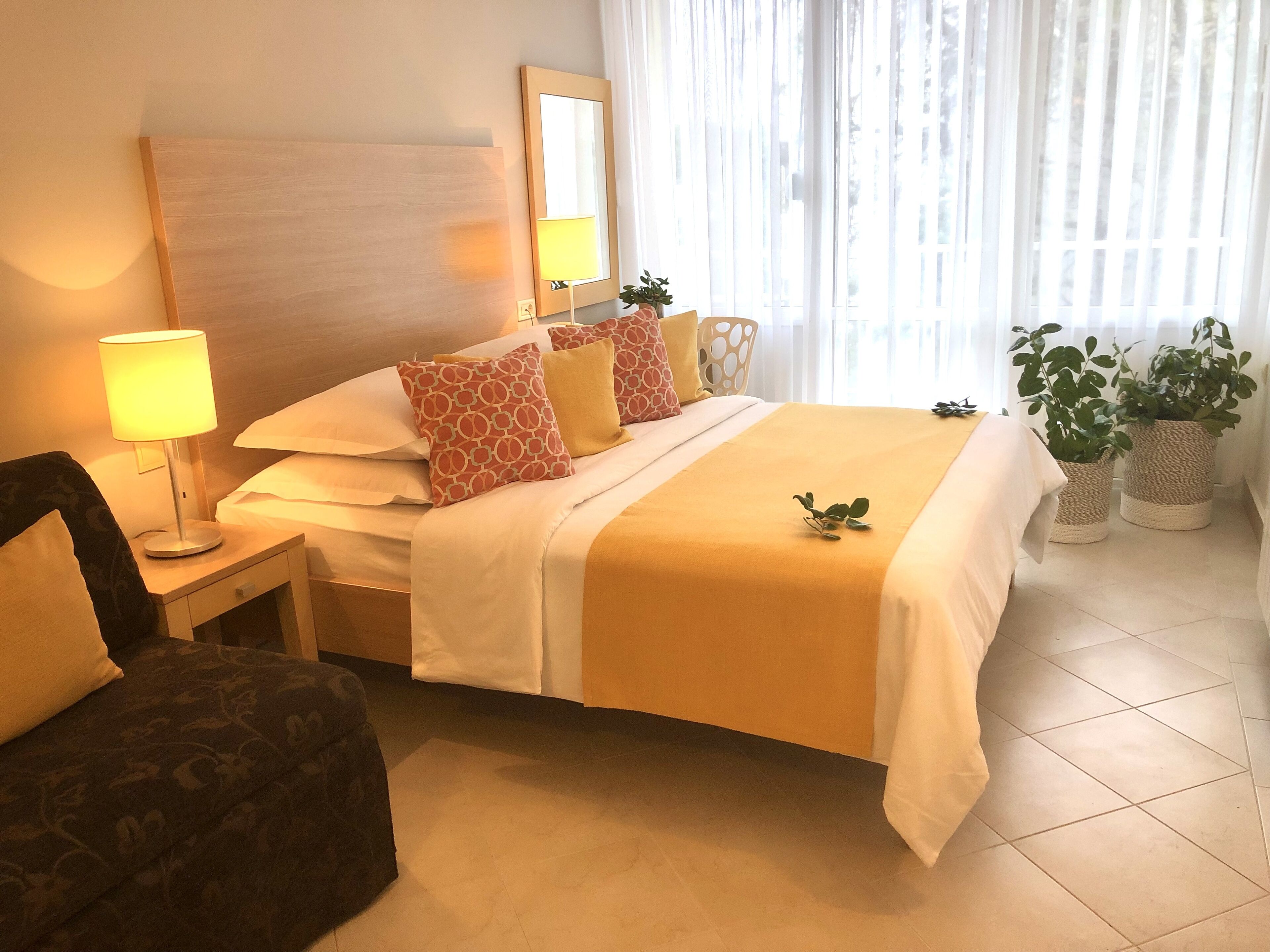 A hotel room with a bed, a nightstand with a lamp, a chair, and two potted plants.