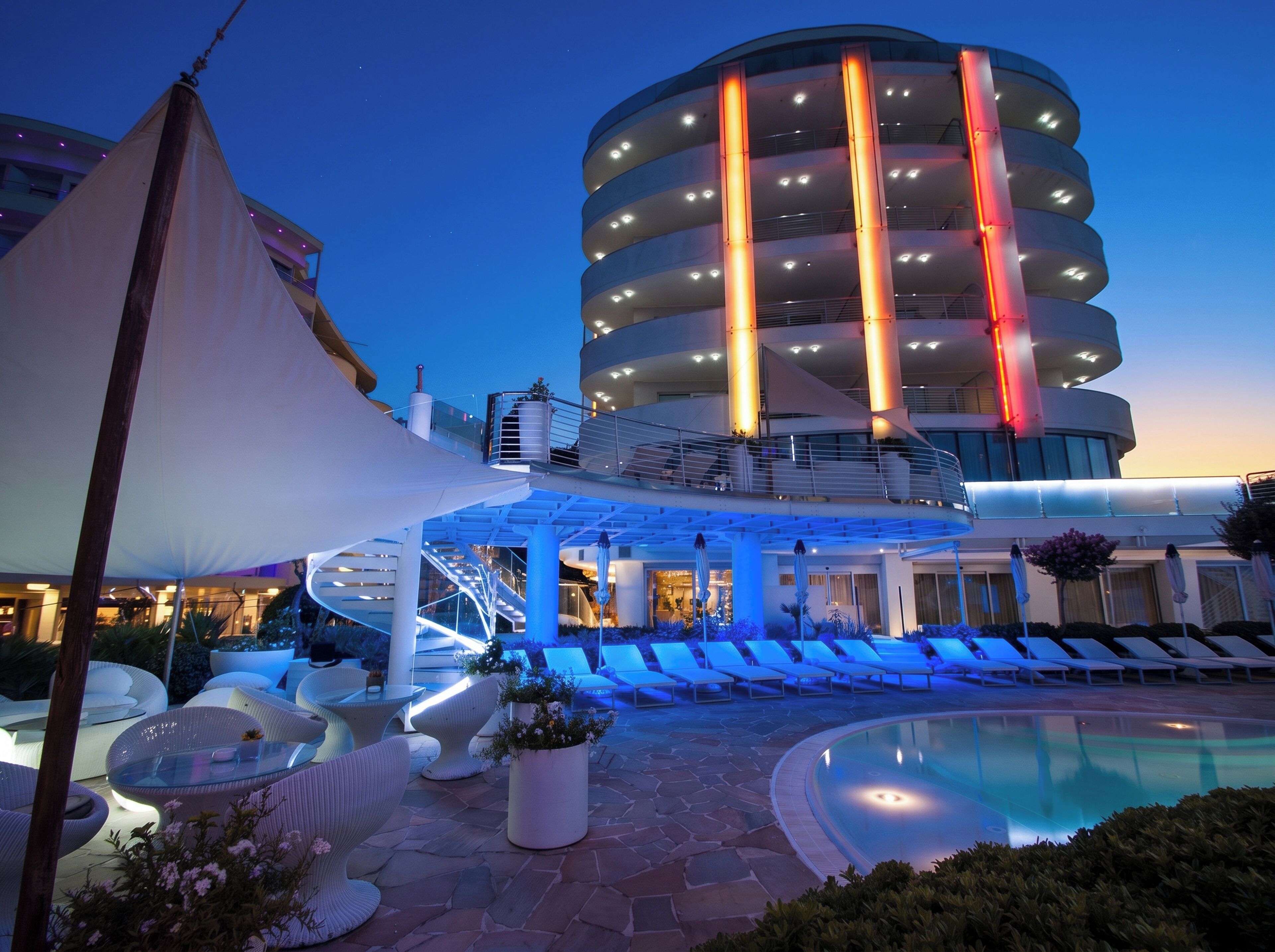 Ein modernes Hotel mit Pool, Liegestühlen und einem markanten zylindrischen Gebäude mit beleuchteten Säulen.