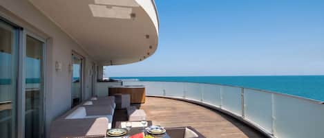 A sunlit outdoor dining area with a view of the sea, featuring a table set with plates, glasses, and a fruit bowl.