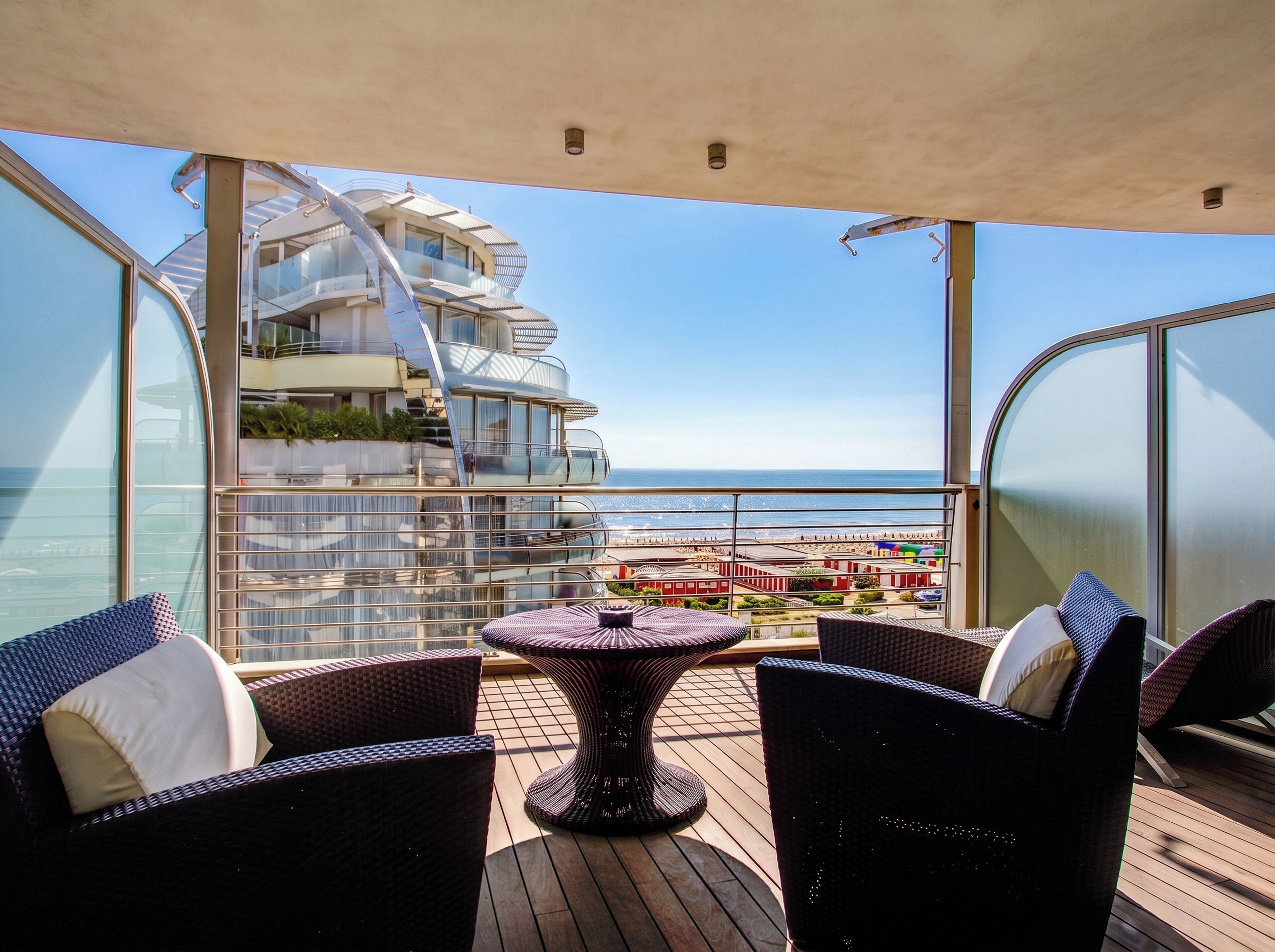 Un balcón con mobiliario de mimbre, barandilla de vidrio y vista a un edificio moderno y al mar.