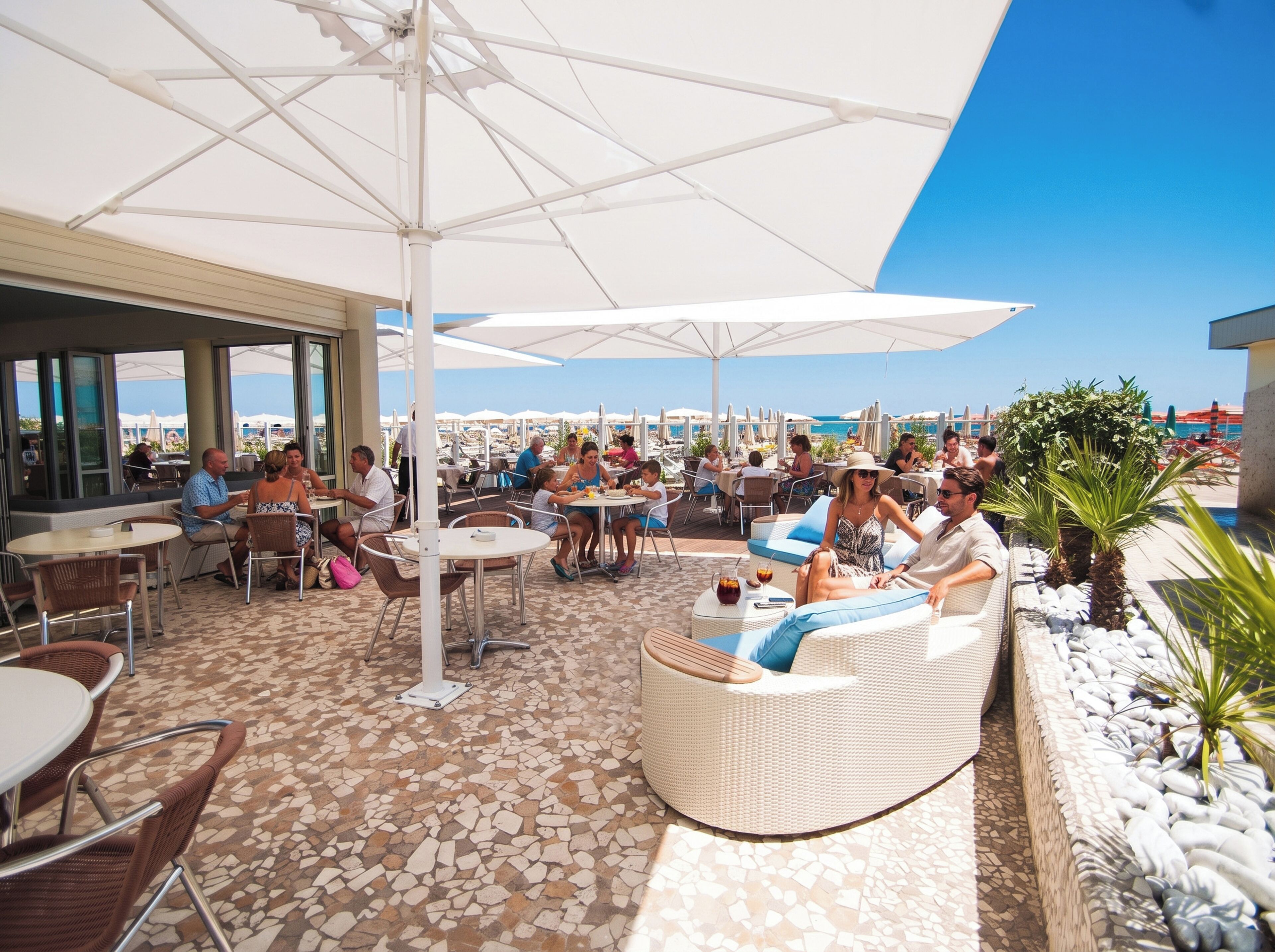 Een terras met tafels en parasols, met uitzicht op een strand.