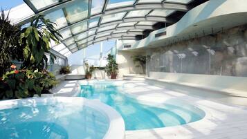 Una piscina interior con techo de cristal, rodeada de plantas en macetas y suelo de baldosas blancas.