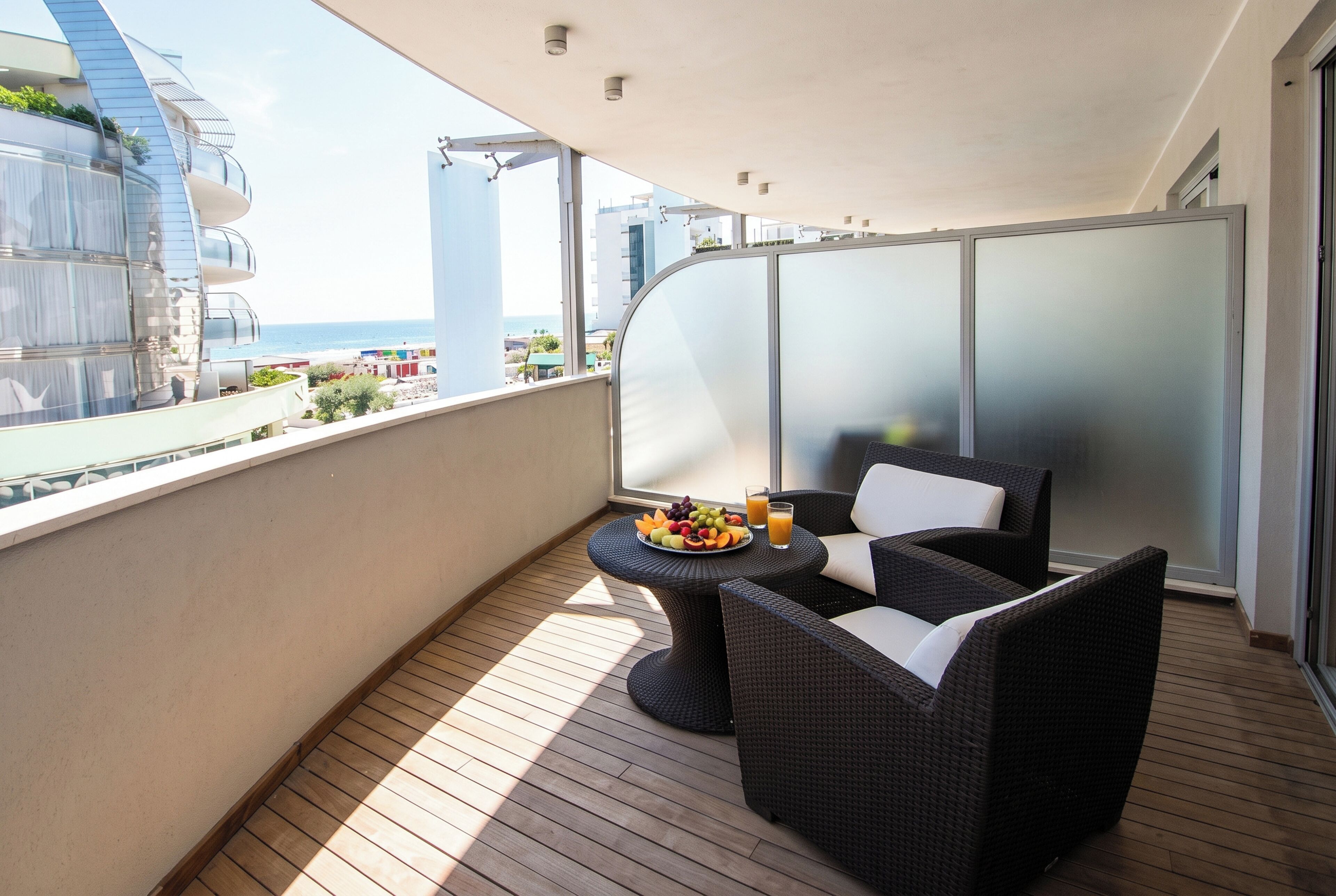 Un balcón con mobiliario de mimbre, una mesa con fruta y jugo, y vistas al mar y edificios.