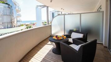 Un balcón con mobiliario de mimbre, una mesa con fruta y jugo, y vistas al mar y edificios.
