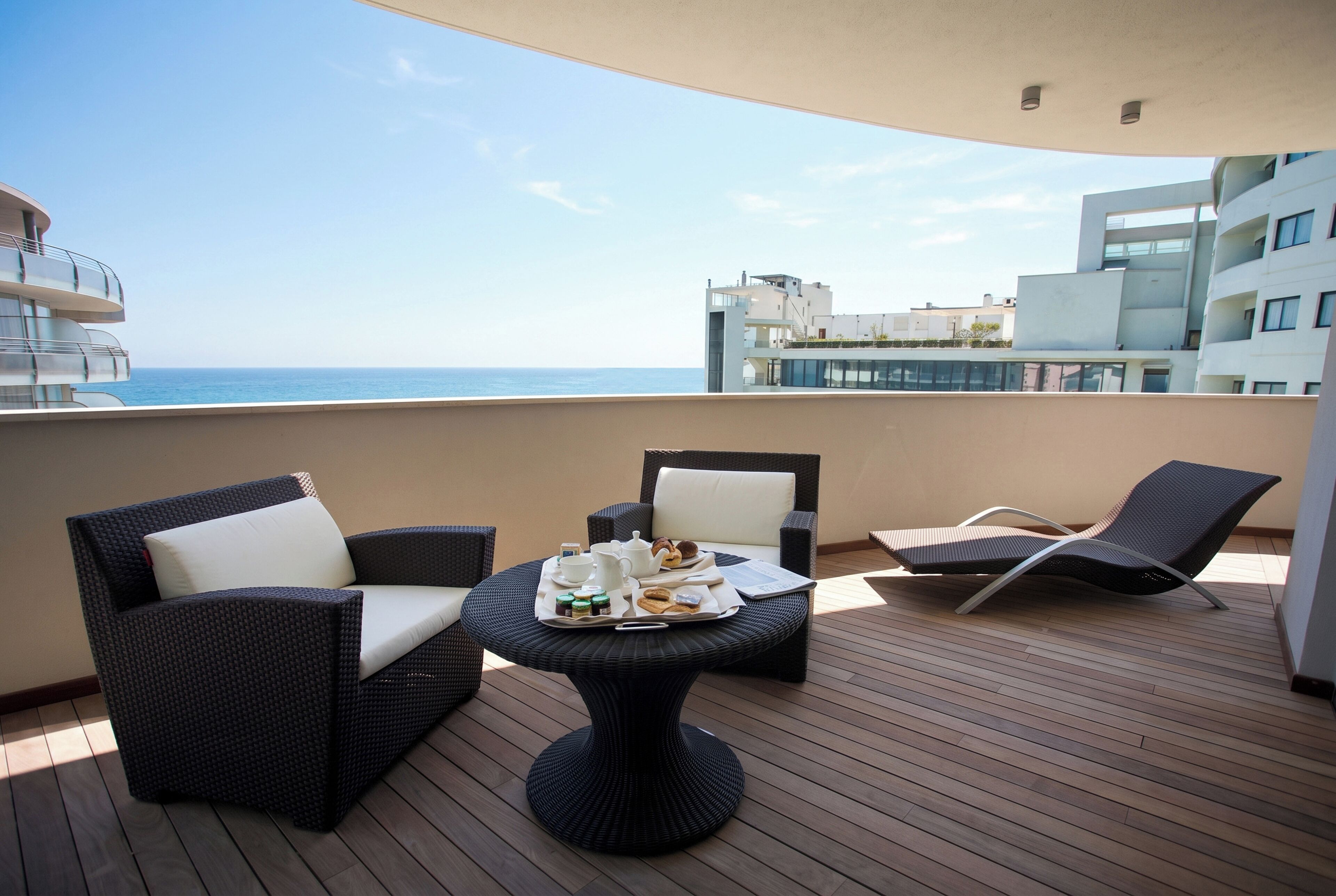 Un balcón con mobiliario de mimbre, una mesa dispuesta con comida y vistas al mar y edificios.