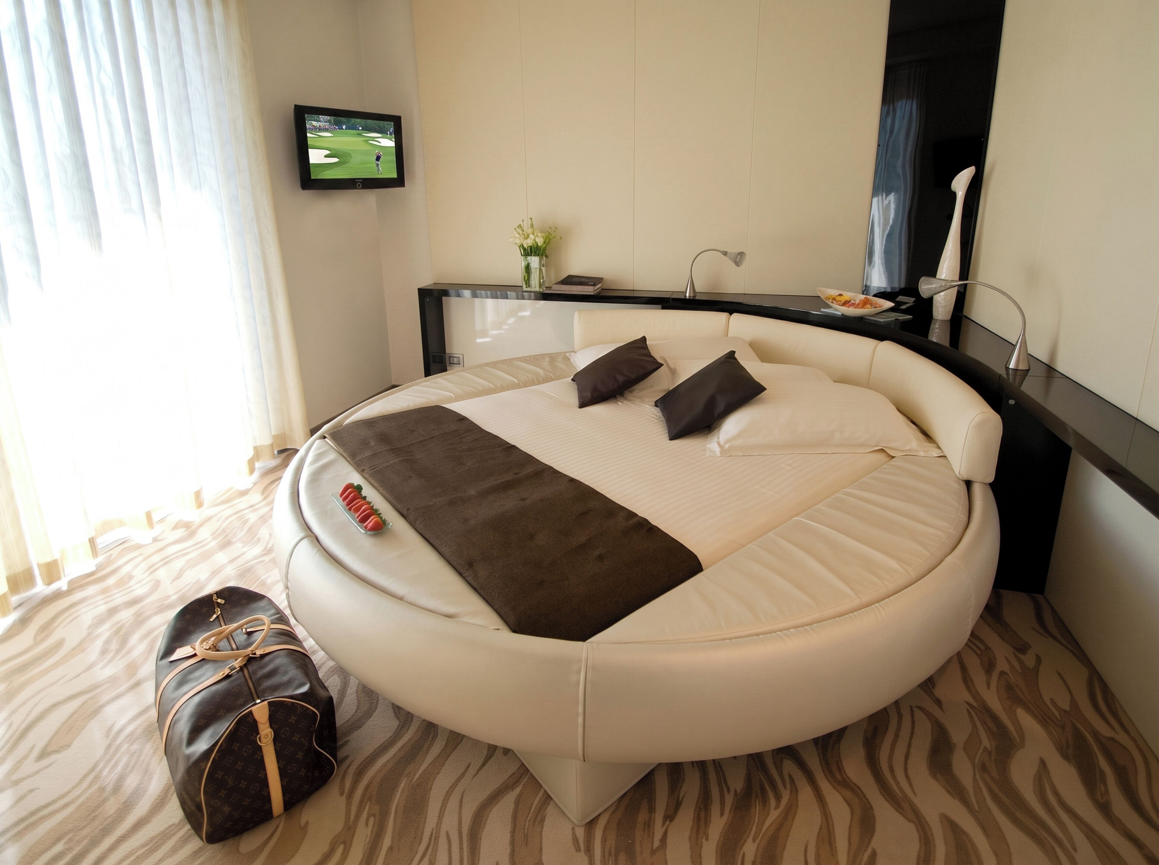 Una habitación de hotel moderna con una cama redonda, un televisor de pantalla plana, una mesita de noche con un jarrón de flores y un bolso de cuero en el suelo.