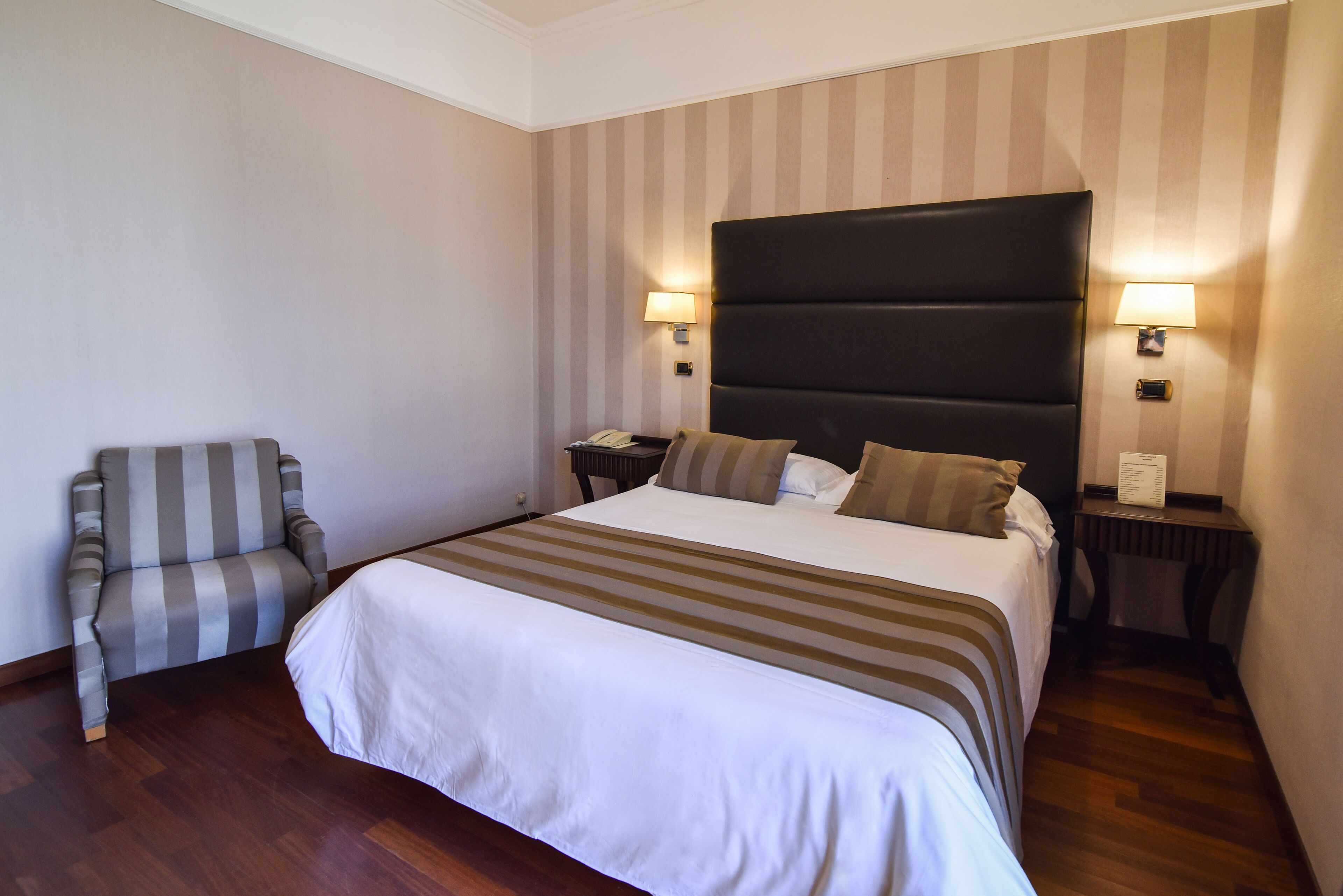 Una camera d'albergo con un letto grande, una testiera a righe, due comodini con lampade, una poltrona a righe e un pavimento in legno.
