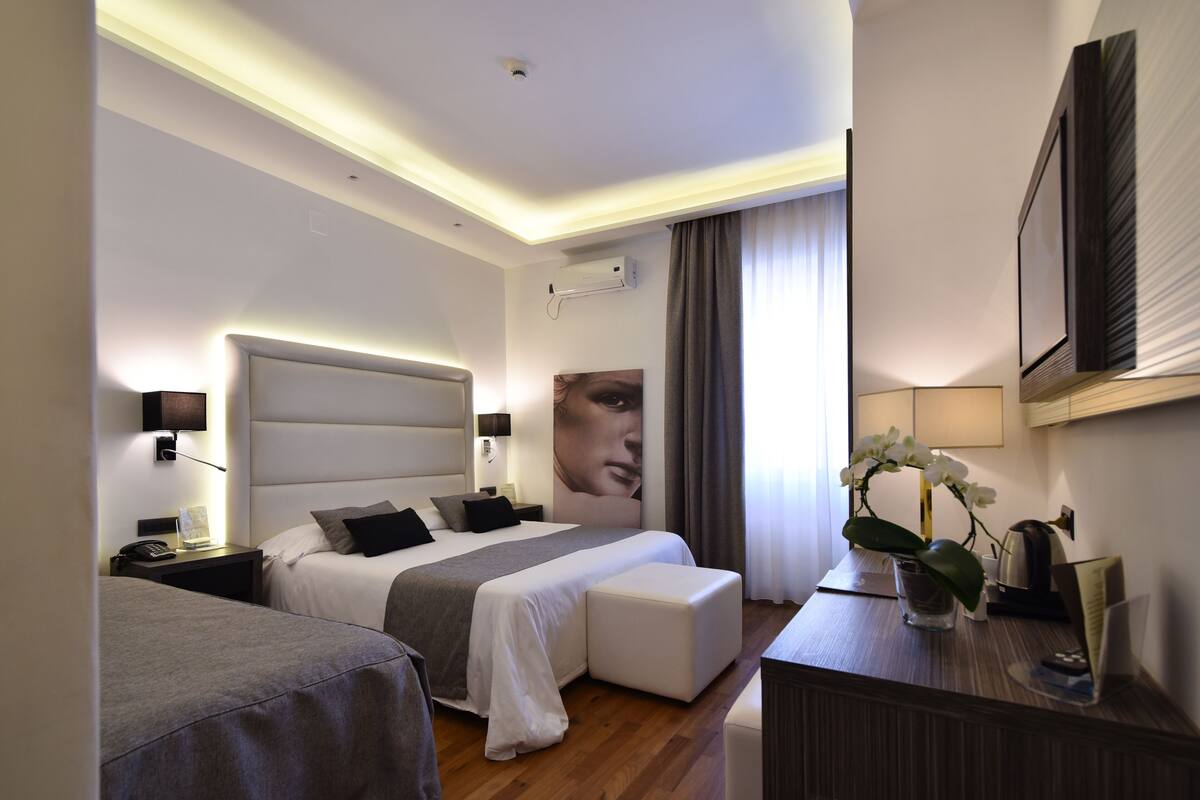 A modern hotel room with a large bed, a smaller bed, a desk with a lamp, and a wall-mounted TV.