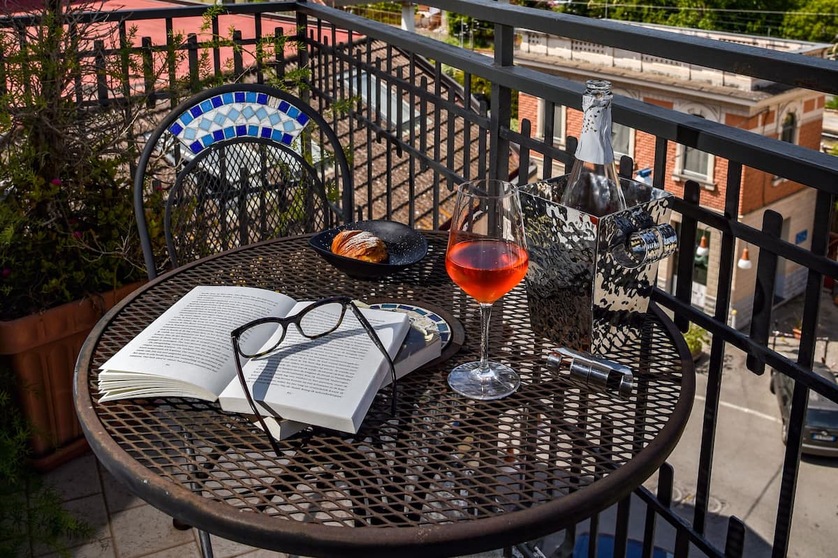 A balcony with a table set for two, featuring a book, glasses, and a bottle.