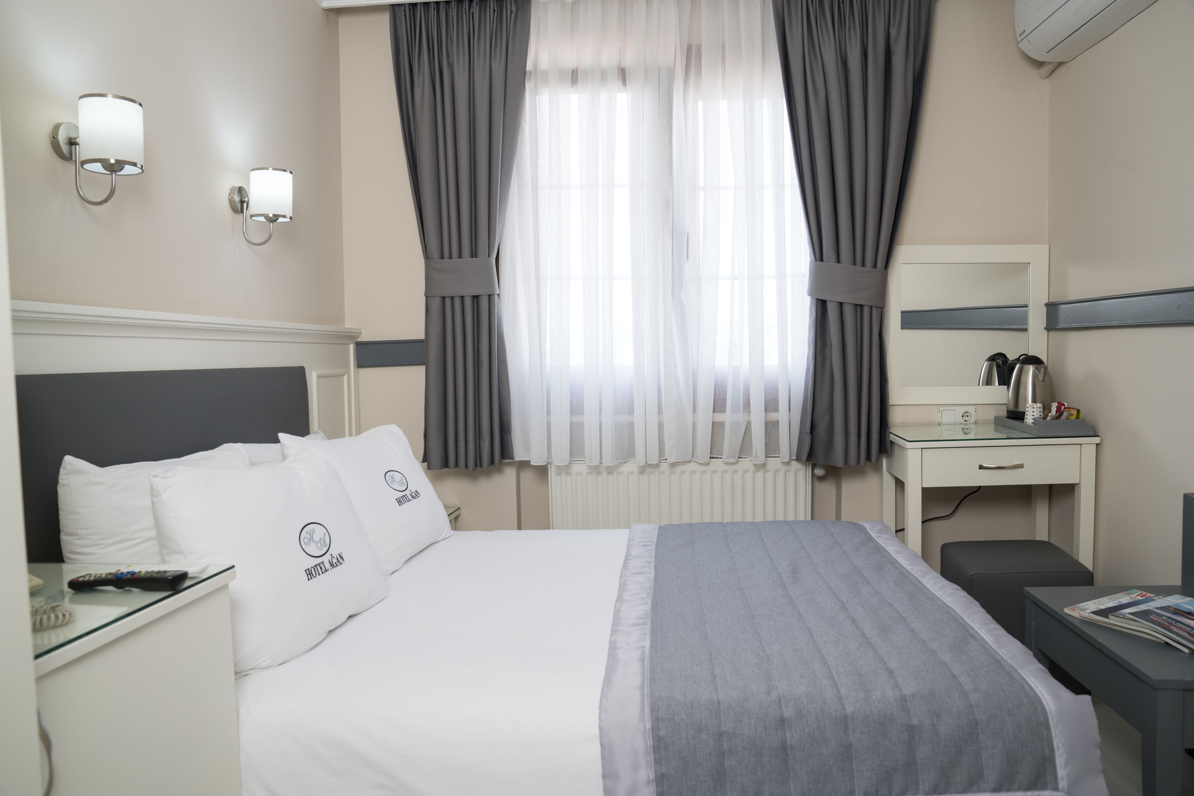 Ein Hotelzimmer mit einem Bett, einem Nachttisch mit Fernbedienung, einem Schreibtisch mit Spiegel, einem Sessel und einem Fenster mit Vorhängen.