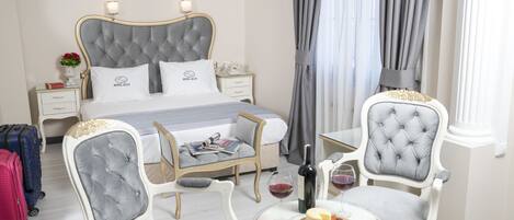 A hotel room with a bed, two armchairs, a small table with a bottle of wine and cheese, and a nightstand with a lamp and a vase of flowers.