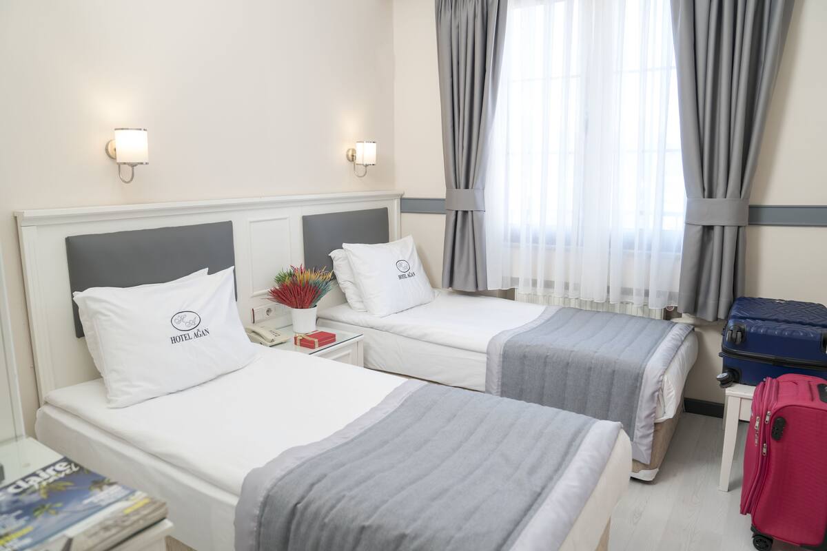 Habitación de hotel con dos camas, una mesita de noche, una lámpara, una maleta y un ventanal con cortinas.