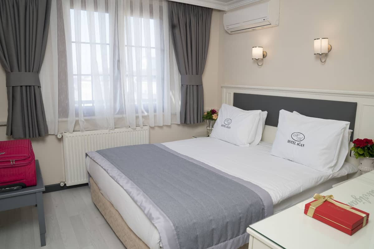 Habitación de hotel con cama, una mesita de noche con una caja de regalo roja, una ventana con cortinas translúcidas y un aire acondicionado de pared.