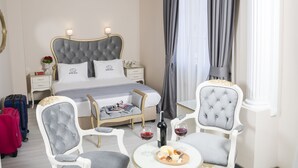 Une chambre d’hôtel avec un grand lit, deux tables de chevet, une petite table de nuit, une chaise, une petite table avec des verres à vin et une bouteille, et une valise.