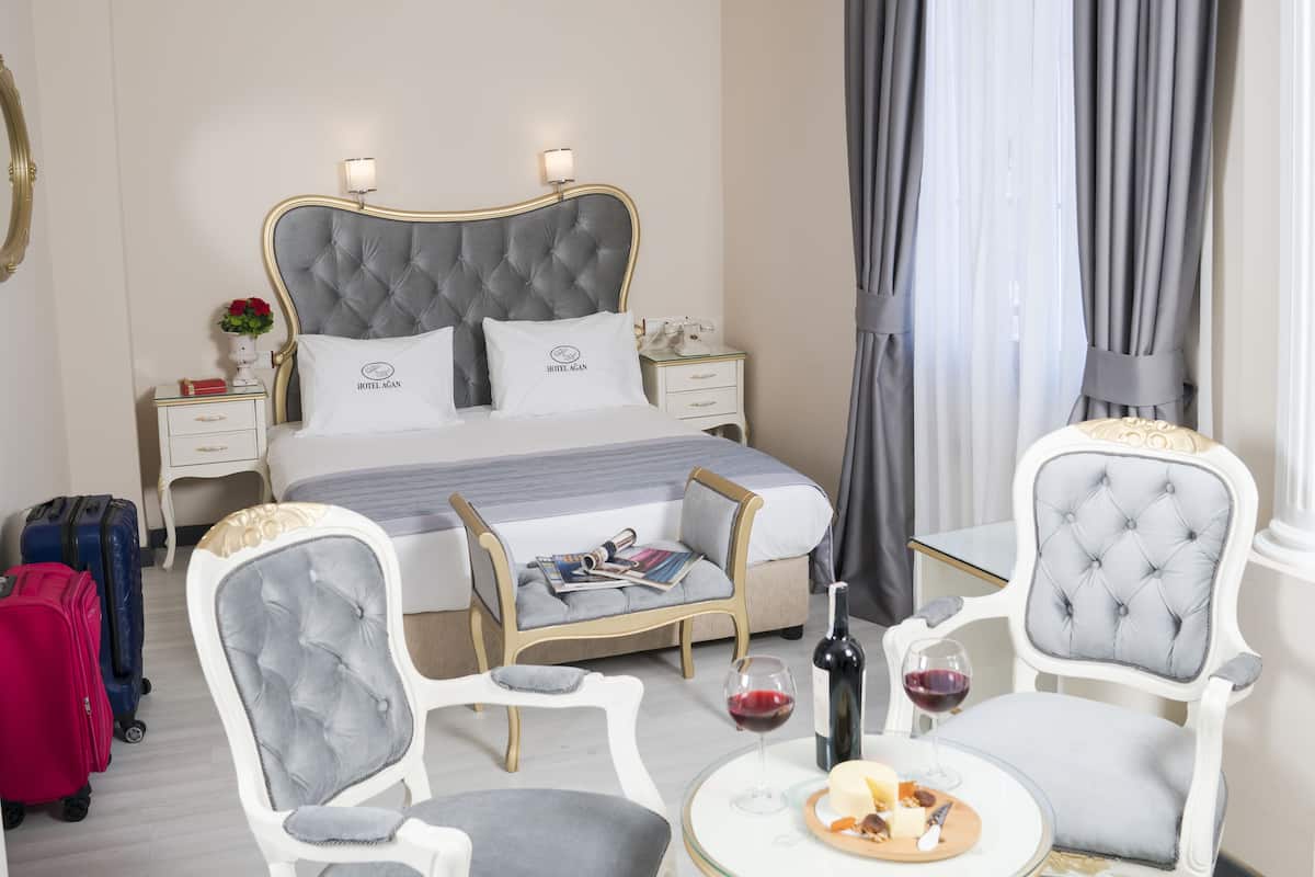 Una habitación de hotel con una cama grande, dos mesitas de noche, una silla, una pequeña mesa con copas de vino y una botella, y una maleta.