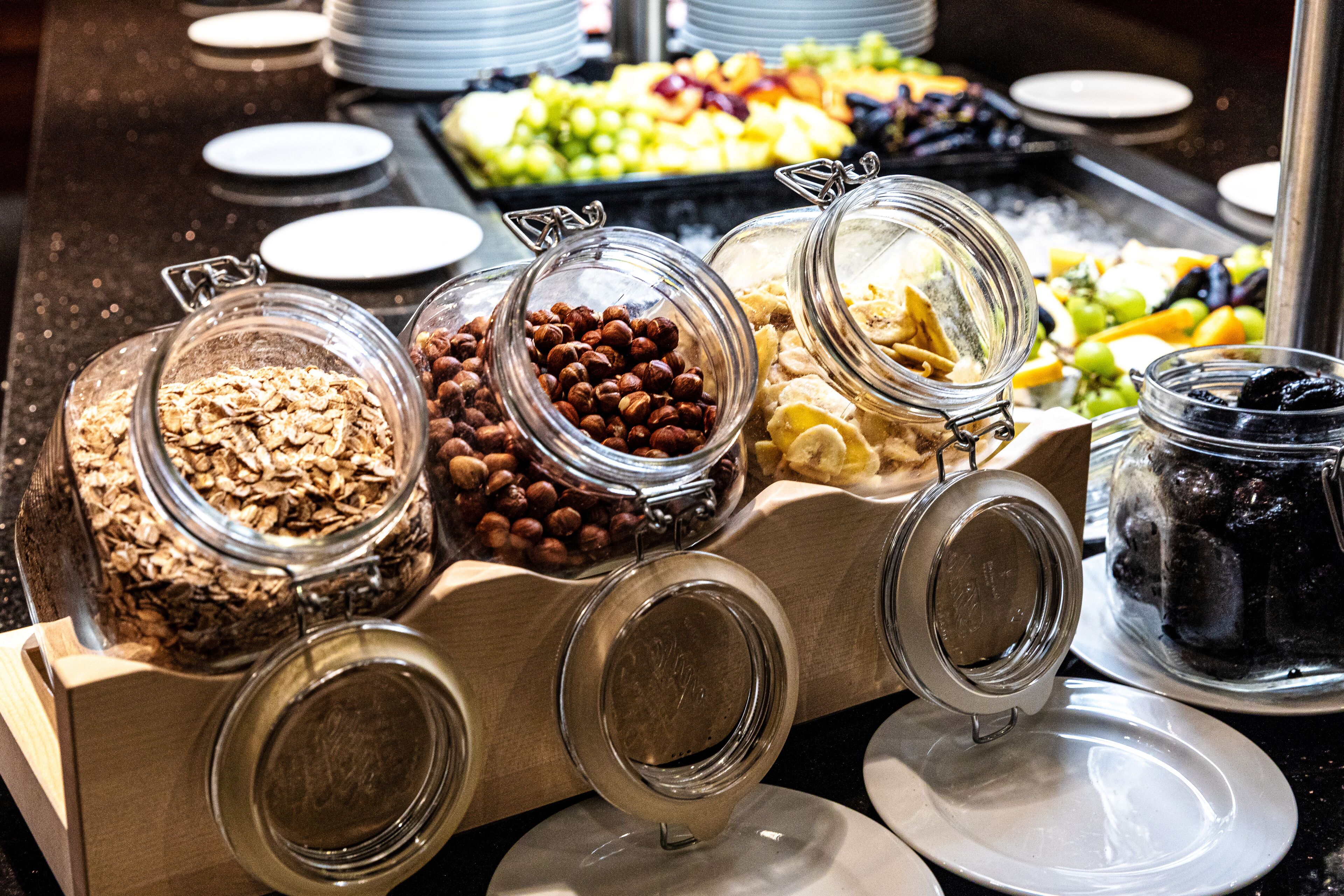 Un bufé con diversos alimentos en recipientes de vidrio, como cereales, frutos secos y frutas.