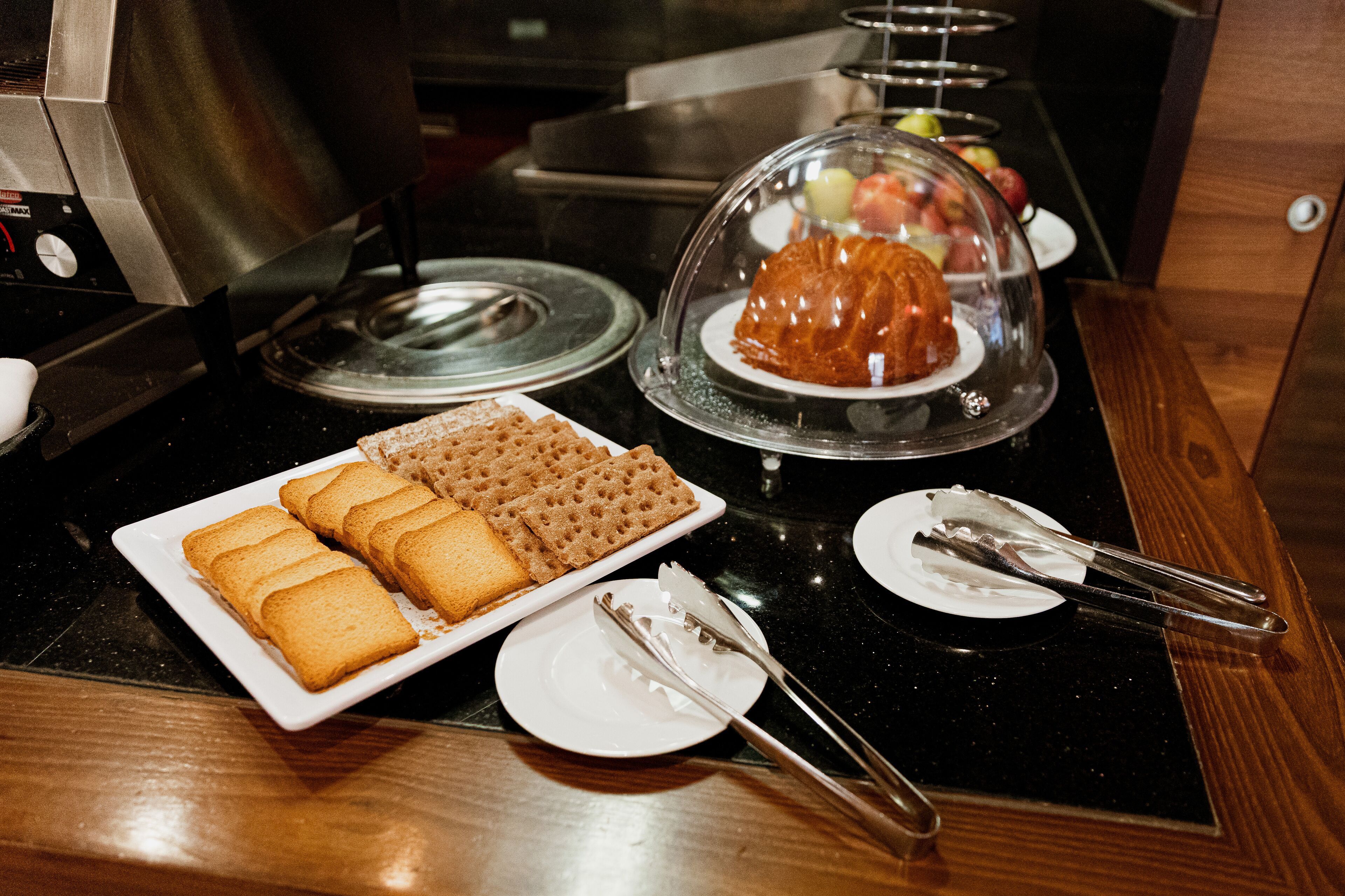 Una mesa de desayuno buffet con pan, un postre cubierto y cubiertos sobre una encimera negra.