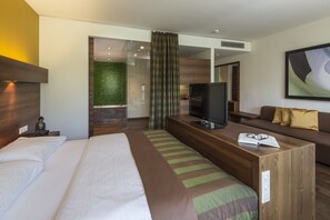 A modern hotel room with a large bed, a wooden desk with a TV, a sofa, and a green-striped ottoman. - Hotel & Asia Spa Leoben (Leoben)