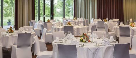 Un salón de banquetes con mesas redondas dispuestas para comer, ventanas grandes con cortinas y vistas a zonas verdes al exterior.
