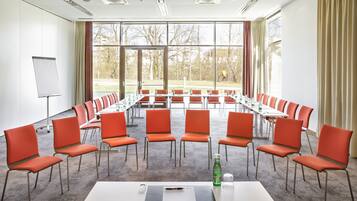 A conference room with a long table, orange chairs, a whiteboard, and large windows with a view of trees.