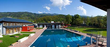 A swimming pool with a clear view of the surrounding area, including a building with a red section and a grassy lawn with lounge chairs.