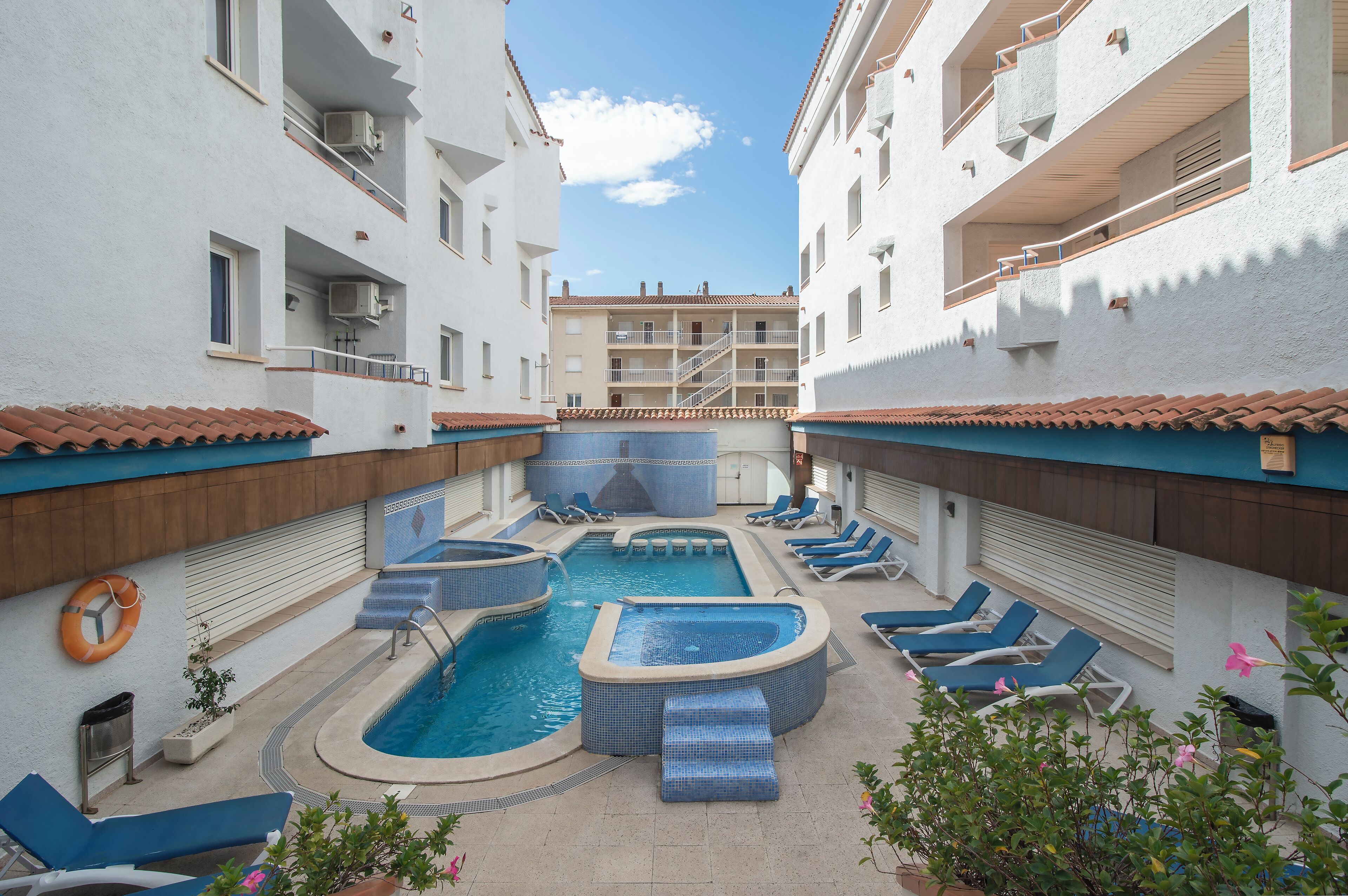 Un área de piscina con varios jacuzzis y sillones de descanso, rodeada de edificios blancos con balcones.