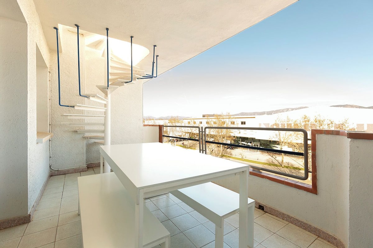Een balkon met een witte tafel en stoelen, uitzicht op een verafgelegen landschap en een trap naar een hoger gelegen niveau.