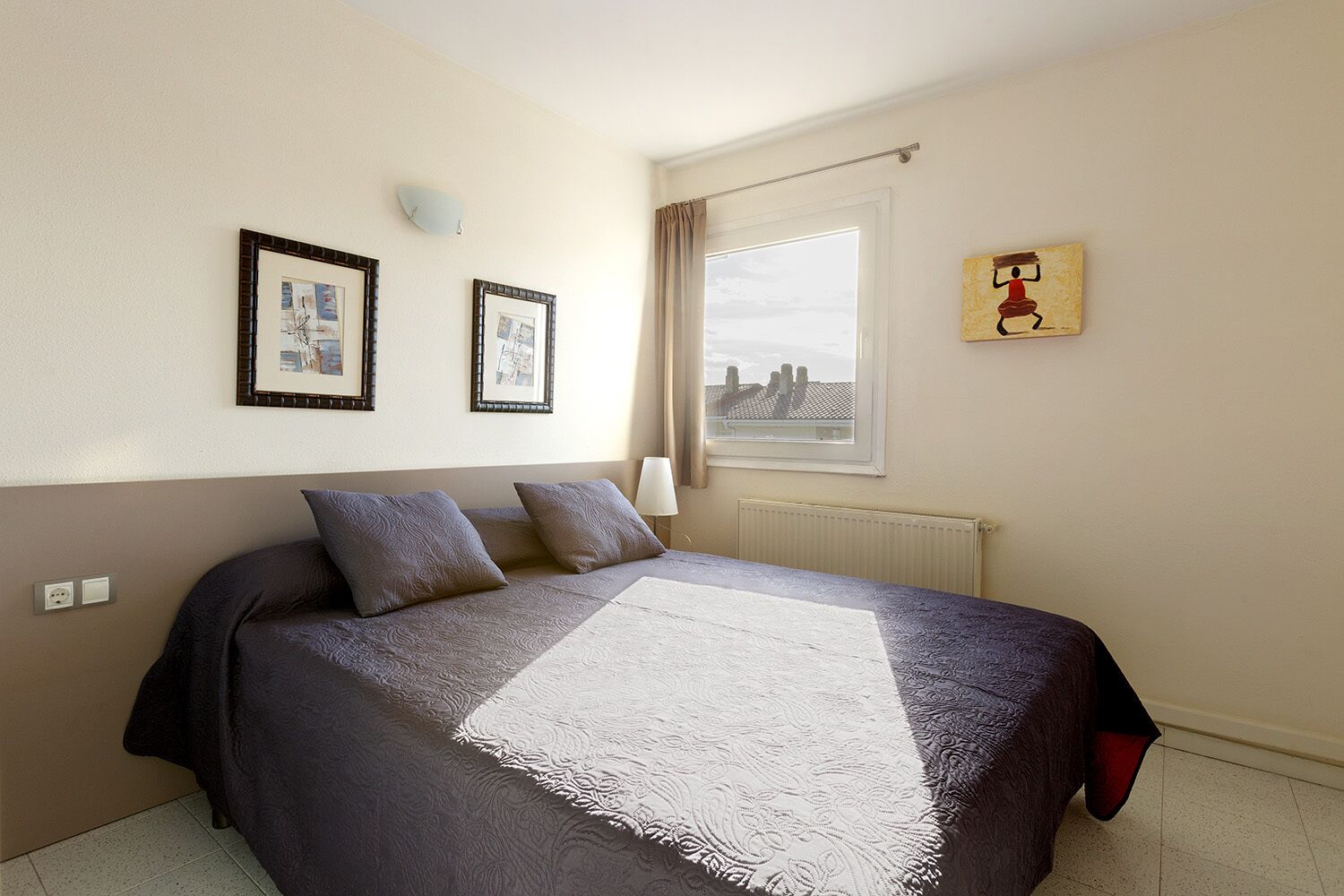 Une chambre à coucher bien rangée, avec un grand lit, deux tableaux encadrés au mur, une fenêtre avec des rideaux et un radiateur.