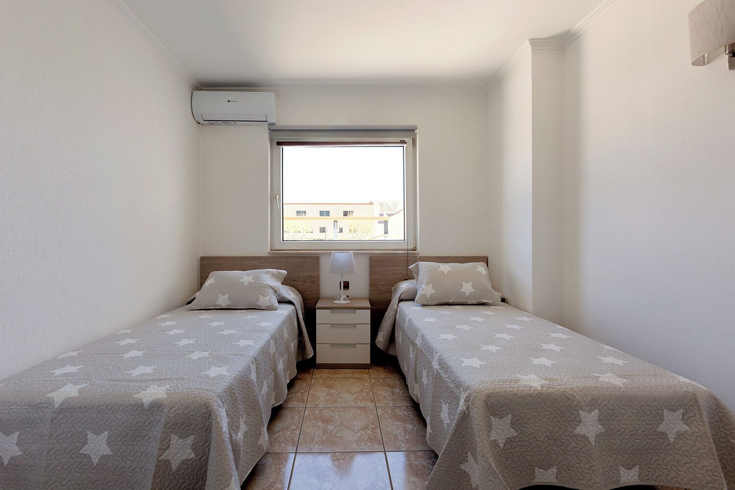 Une petite chambre propre, avec deux lits, une table de chevet, une fenêtre et un appareil de climatisation.