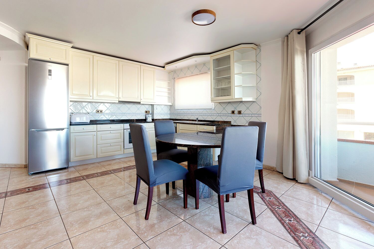 A modern kitchen with a built-in refrigerator, white cabinets, a dining table with chairs, and a large window with a view of the outside.