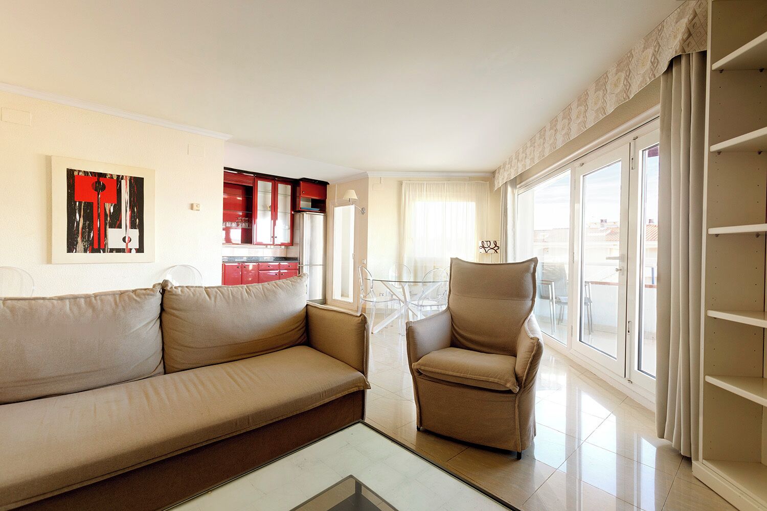 A living room with a beige sofa and armchair, a glass coffee table, and a red kitchen in the background.