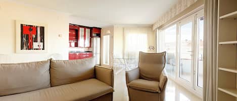 A living room with a beige sofa and armchair, a glass coffee table, and a red kitchen in the background.