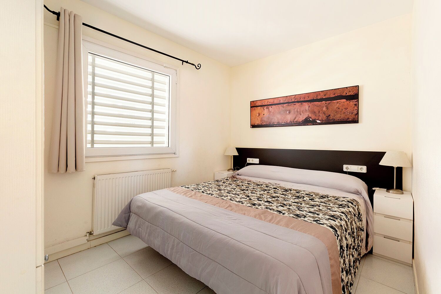 A bedroom with a bed, a radiator, a window with curtains, a wall-mounted artwork, and bedside lamps.
