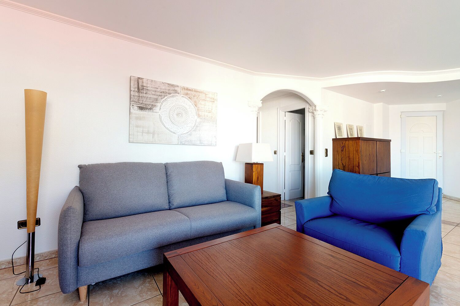 Una sala de estar con un sofá gris, un sofá azul, una mesa de centro de madera, una lámpara y un armario.