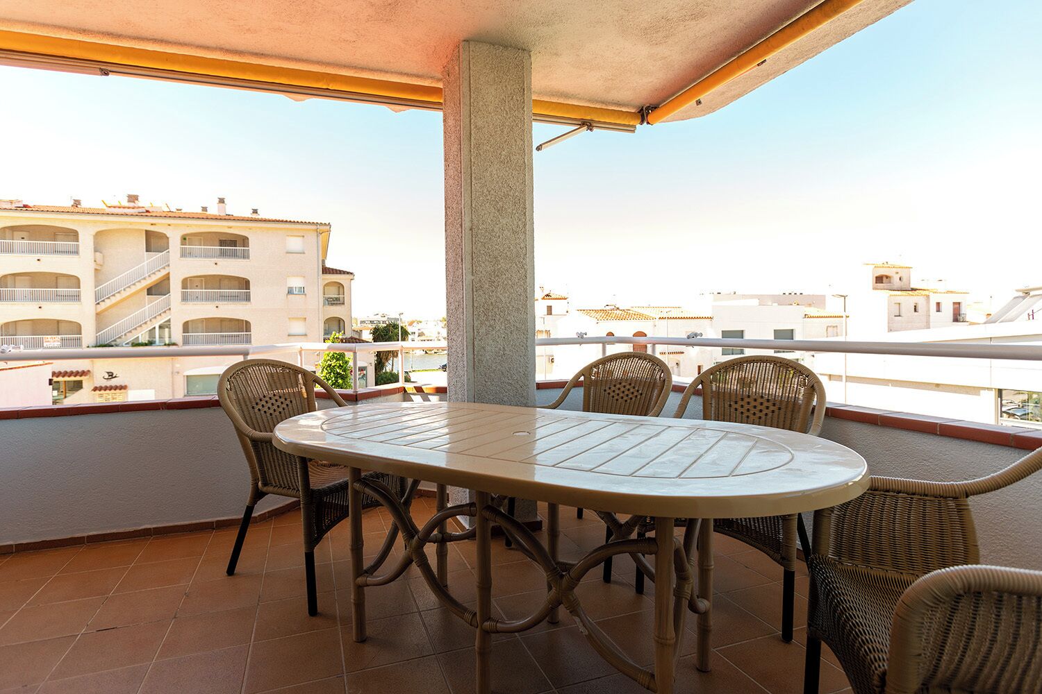 Un balcón con una mesa redonda y sillas de mimbre, con vistas a una zona residencial con edificios de varios pisos.